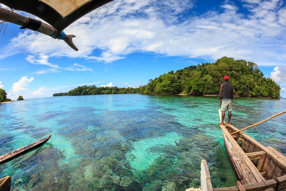 A Titan villager navigates a traditional melanesian outrigger over shallow corals that flank a small island in the Manus Province of Papua New Guinea. Mystery shrouds the densely vegetated island, as legend amongst the indigenous Mbuke people maintains that it is home to a "dinosaur" that has never been seen on any other island in the Archipeligo.
